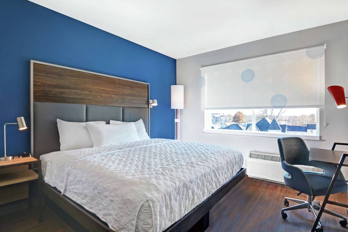 Une chambre d’hôtel moderne avec un grand lit, un bureau avec une chaise, une fenêtre avec des stores et une lampe sur la table de chevet.