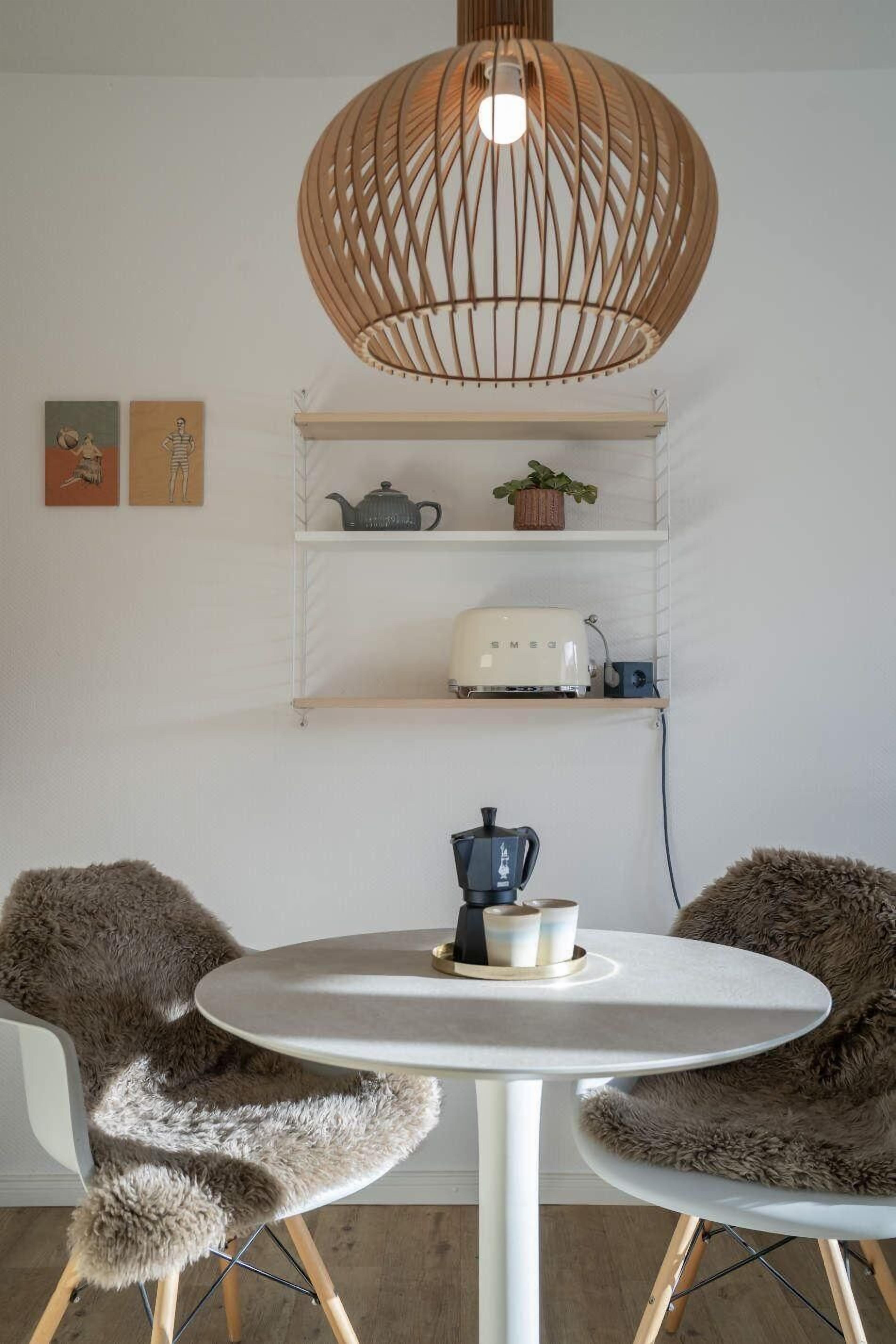 Ett modernt kök med ett runt bord, två stolar, en hängande lampa och hyllor med köksredskap.