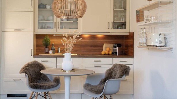 Una cocina moderna con armarios blancos, una mesa de comedor redonda, dos sillas, una lámpara colgante y una pequeña planta en la encimera.