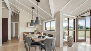 A dining area with a wooden table, grey chairs, pendant lights, and a view of the outdoors through large windows.