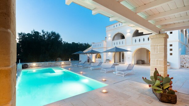 Une piscine entourée de chaises longues et agrémentée de bougies, le tout bordé d’un bâtiment blanc aux arches élégantes.