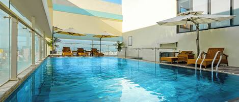 A modern indoor swimming pool with clear blue water, sun loungers, and umbrellas.
