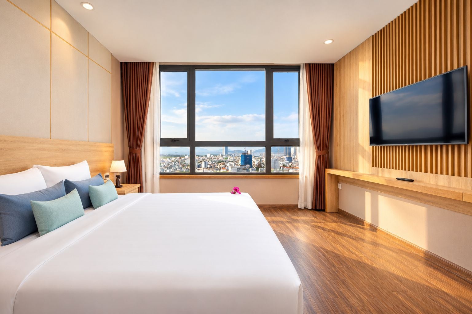 A hotel room with a large bed, a flat-screen TV mounted on the wall, and a city view through the window.