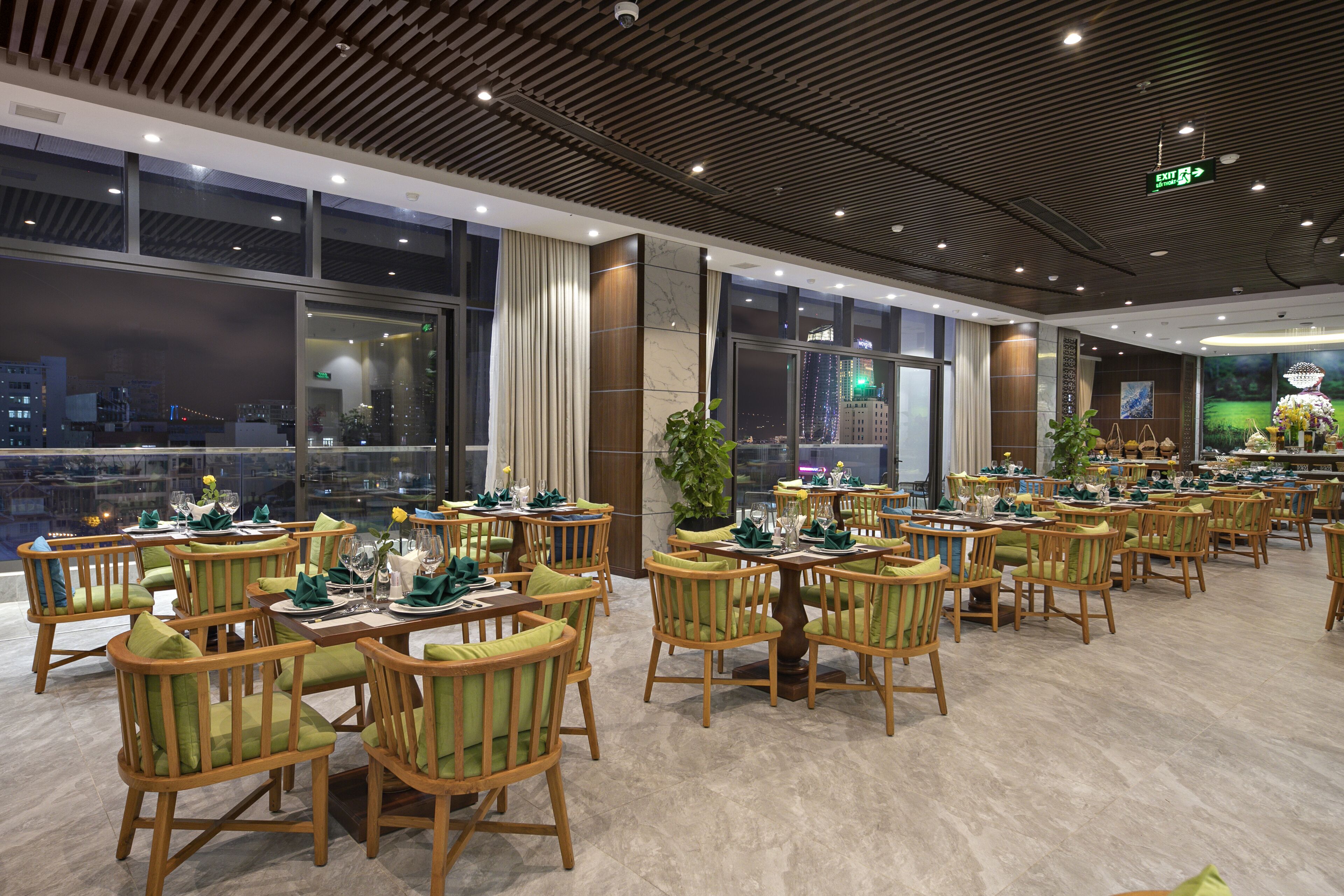 Un restaurante espacioso con sillas y mesas de madera, amplias ventanas que ofrecen vistas a la ciudad y un diseño de techo moderno.
