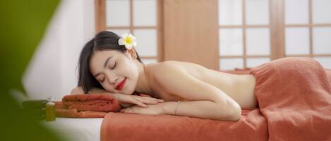 A person lying on a bed with a towel, a bottle, and a flower in a room with a window and wooden paneling.