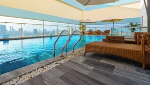 An indoor swimming pool with clear blue water, a city skyline view, and a seating area with wicker furniture and umbrellas.
