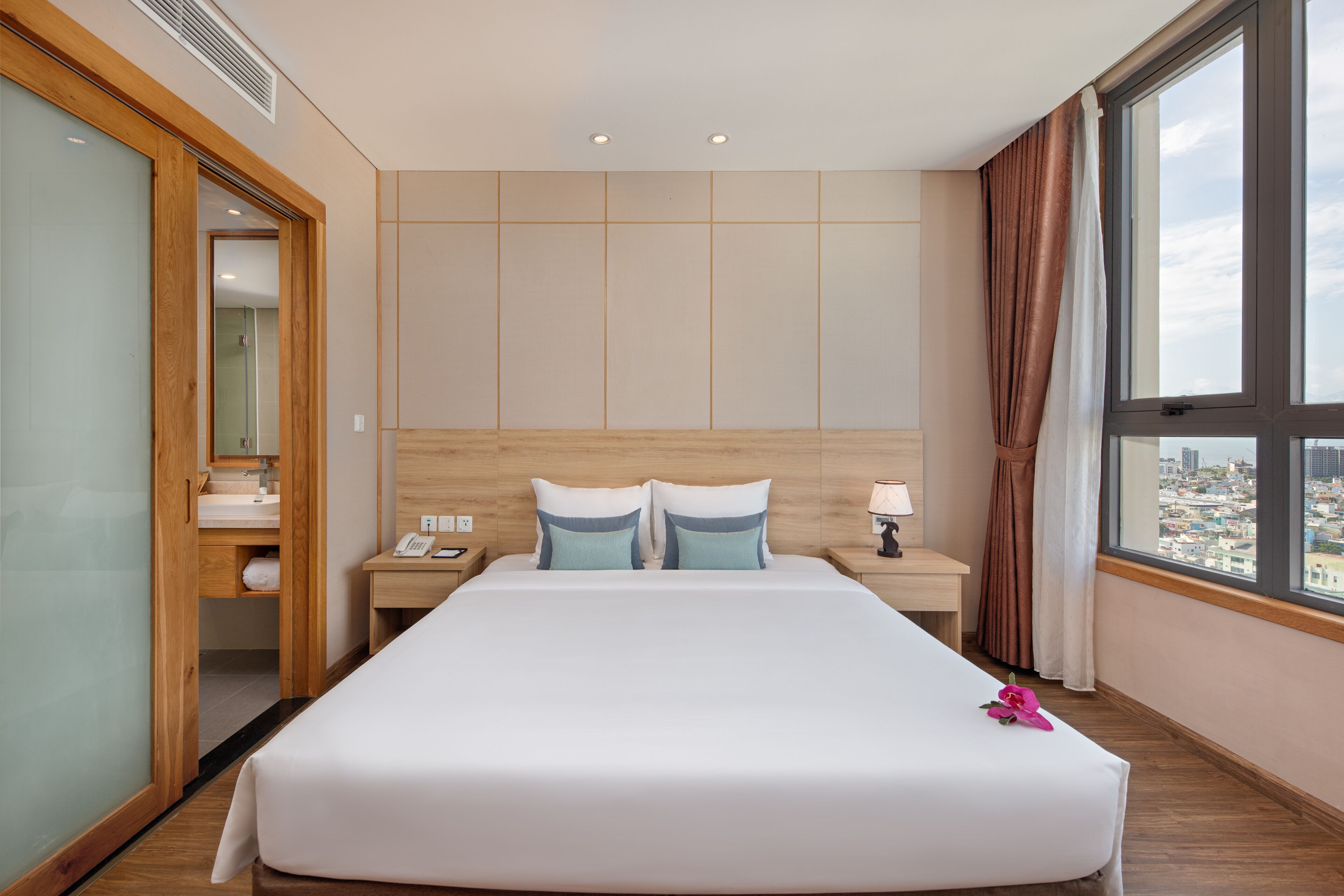 A hotel room with a large bed, wooden headboard, a nightstand with a lamp, a mirror, and a view of the city through a large window.
