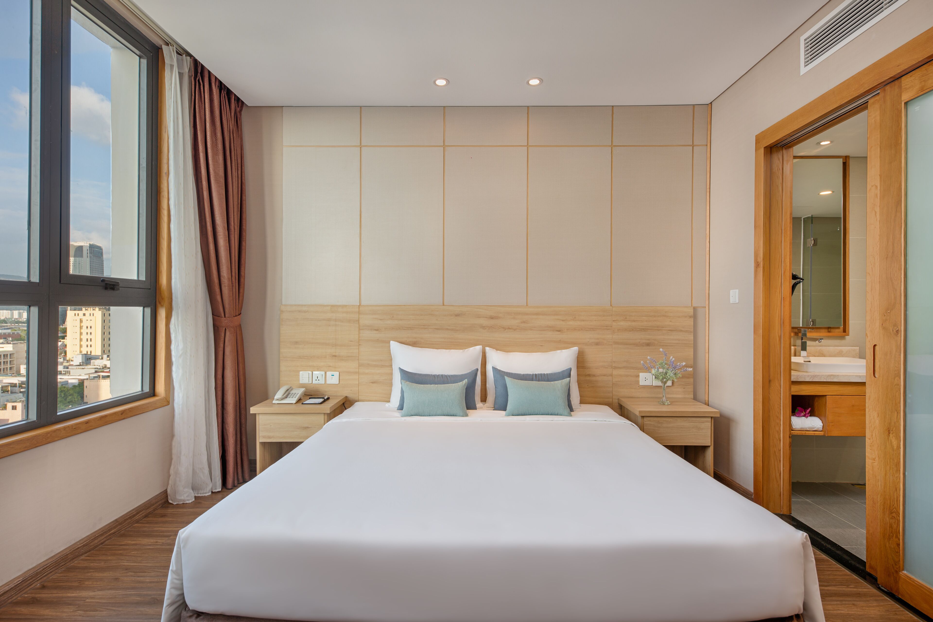 A modern hotel room with a large bed, wooden headboard, a nightstand, a bedside table, a window with curtains, and a view of the city.