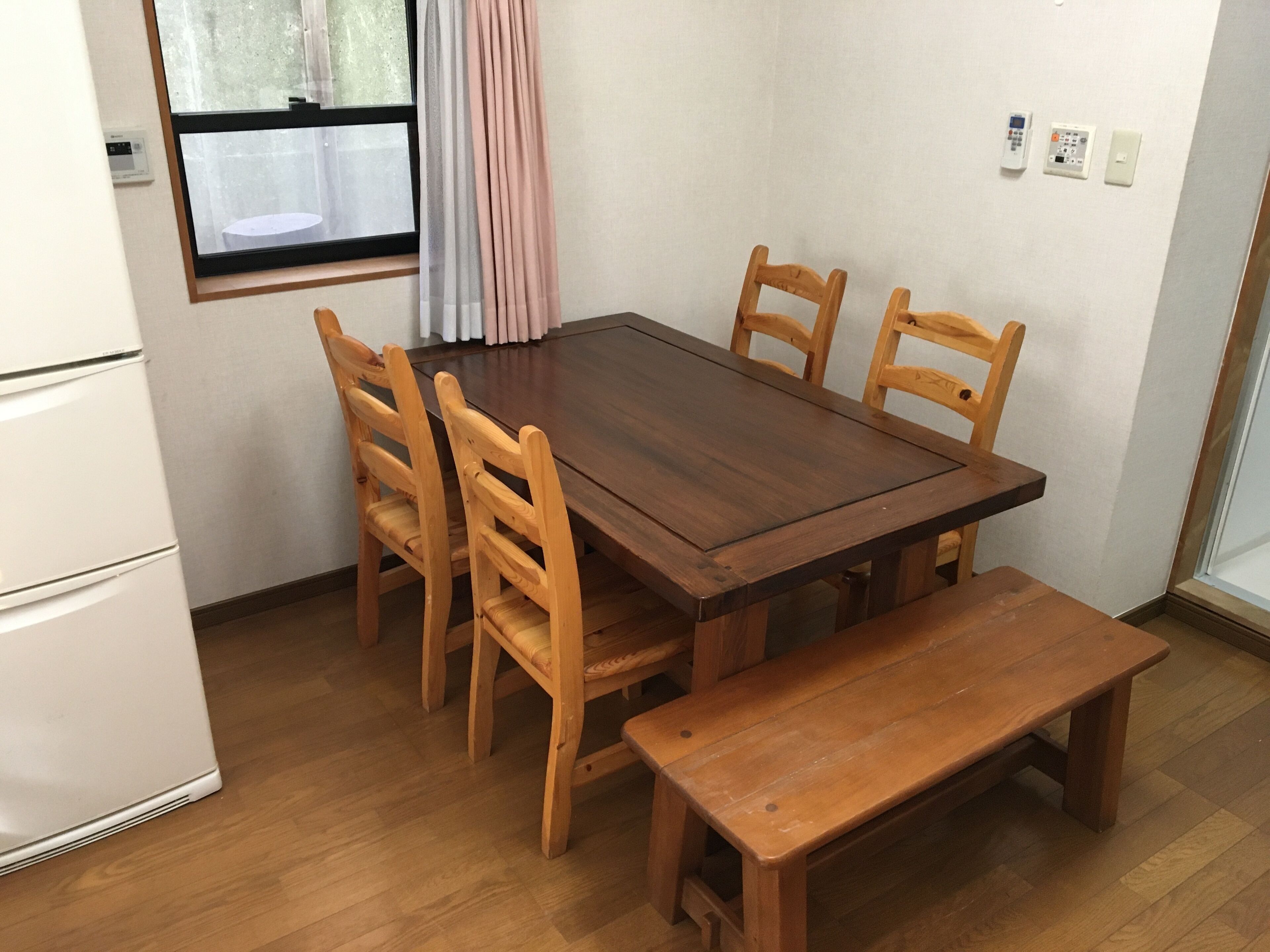 A dining area with a wooden table, six chairs, a bench, a refrigerator, a window with curtains, and a thermostat on the wall.