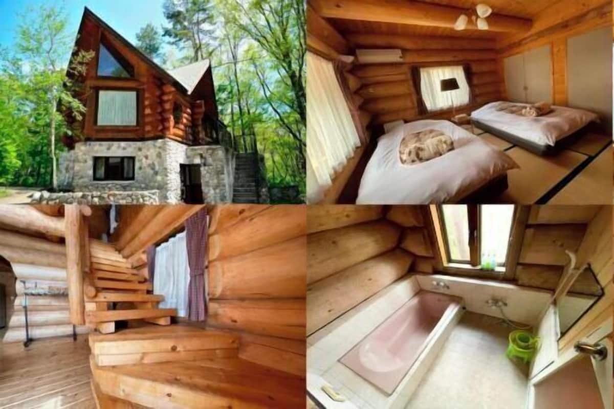 A wooden cabin with a stone foundation, a bedroom with a bed and a window, a dining area with a table and chairs, a bathroom with a bathtub, and a staircase.