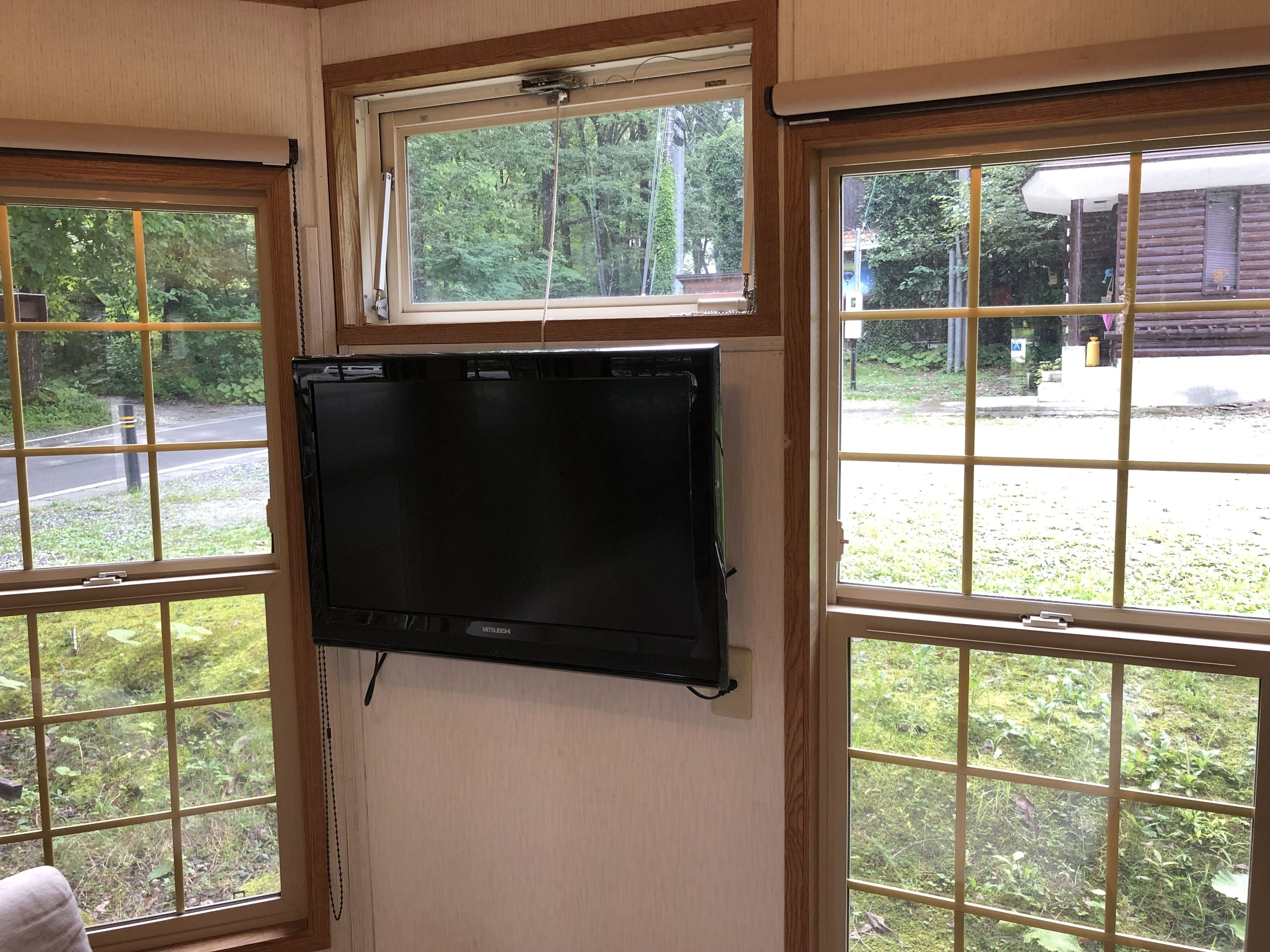 Una habitación con un televisor de pantalla plana negro grande montado en la pared, ventanas de madera con diseño de cuadrícula y vistas a una carretera y vegetación al exterior.