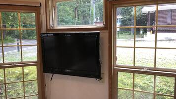 Una habitación con un televisor de pantalla plana negro grande montado en la pared, ventanas de madera con diseño de cuadrícula y vistas a una carretera y vegetación al exterior.