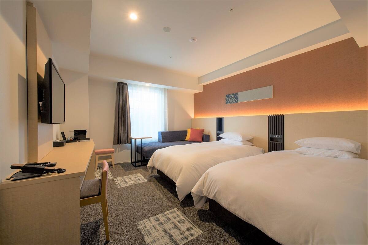 Ein Hotelzimmer mit zwei Betten, einem Schreibtisch, einem Stuhl, einem Fernseher und einem Fenster mit Vorhängen.