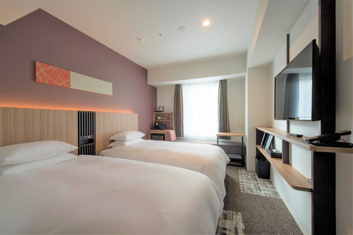 Ein Hotelzimmer mit zwei Betten, einem Fernseher, einem Schreibtisch und einem Fenster mit Vorhängen.