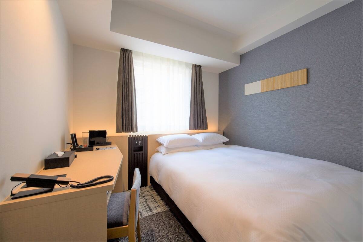 Ein Hotelzimmer mit einem Bett, einem Schreibtisch mit Telefon, einer Lampe und einem Fenster mit Vorhängen.