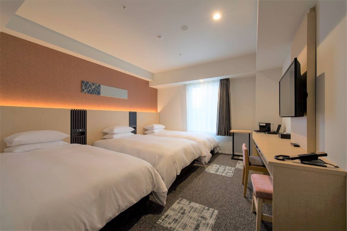 Ein Hotelzimmer mit zwei Betten, einem Schreibtisch, einem Stuhl, einem Fernseher und einem Fenster mit Vorhängen.