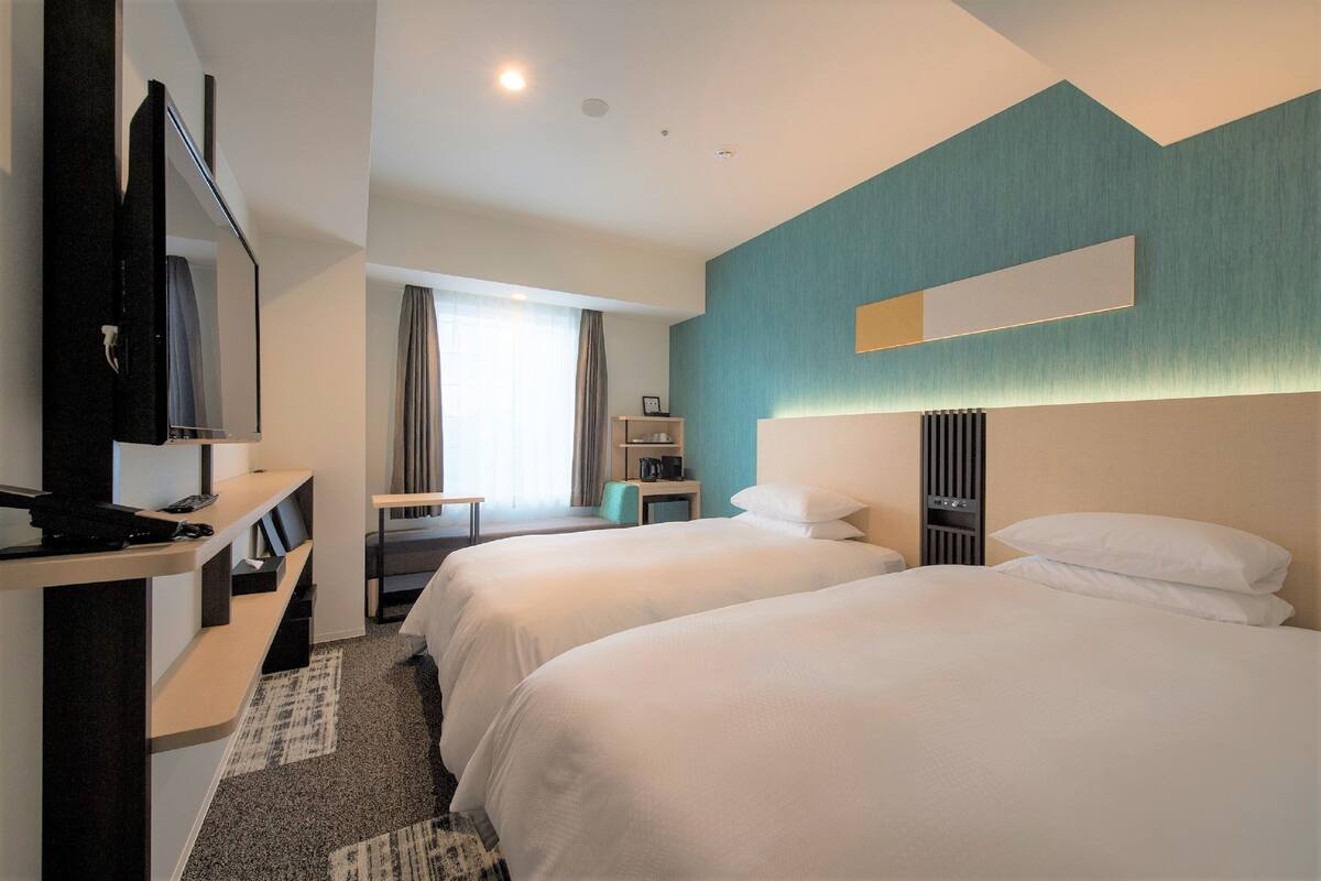 Ein Hotelzimmer mit zwei Betten, einem Schreibtisch, einem Fernseher und einem Fenster mit Vorhängen.