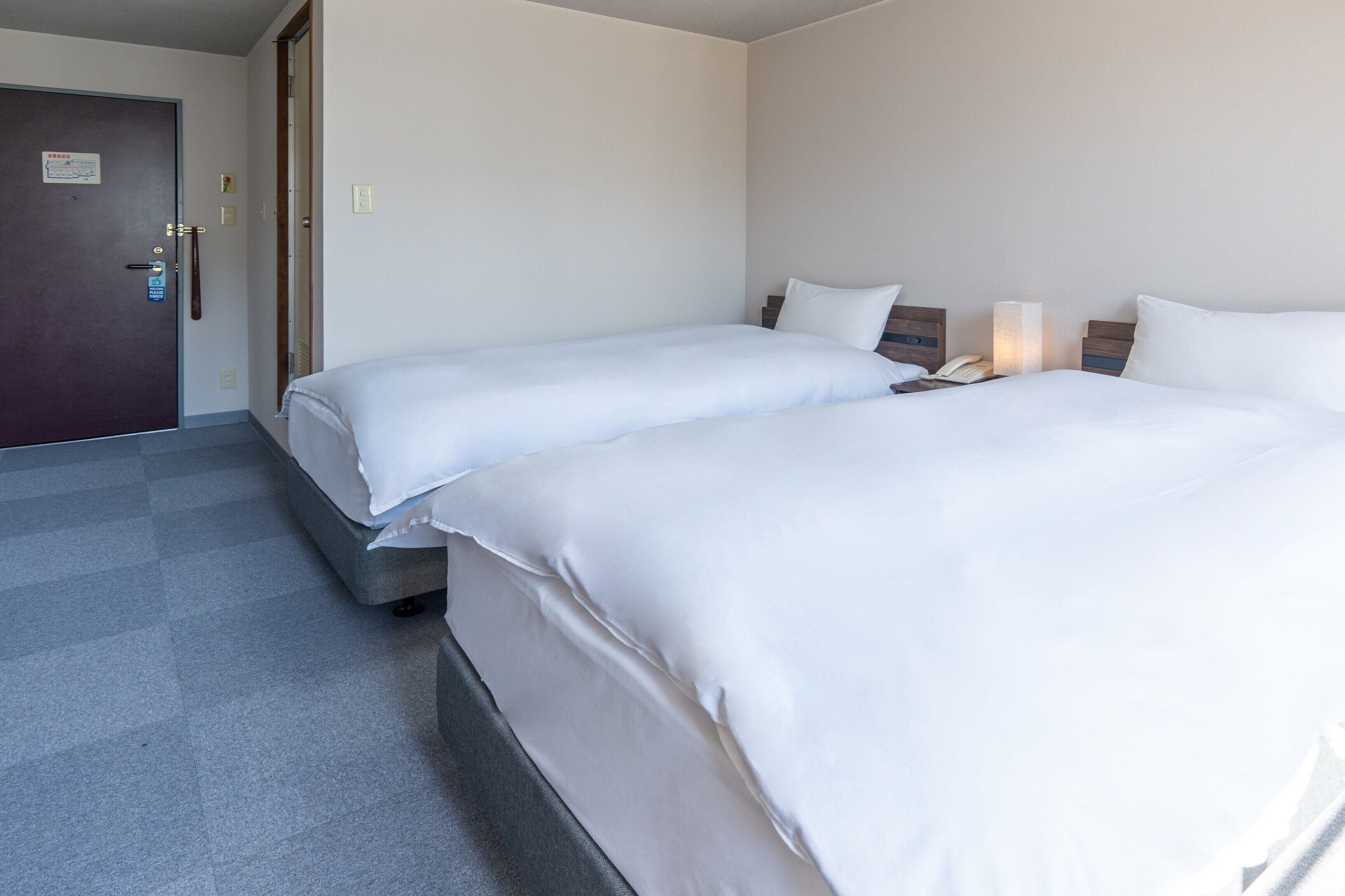 Quarto de hotel com duas camas de solteiro, uma porta com aviso e uma mesa de cabeceira com abajur.