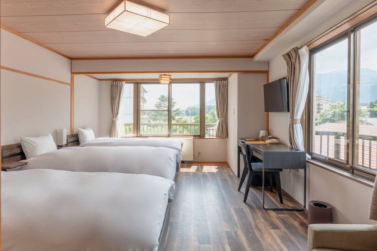 A hotel room with two beds, a desk, a chair, a TV, and large windows with a view of the mountains.