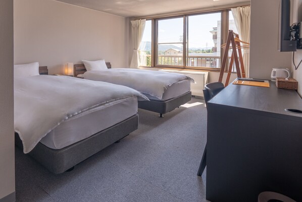 A hotel room with two beds, a desk, a chair, a TV, and a window with a view of buildings and mountains. - SUMMIT VIEWS HAKUBA (Hakuba)