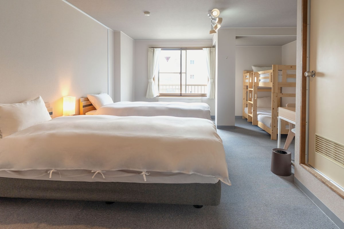 A hotel room with two beds, a window with curtains, a bedside lamp, and a bunk bed in the corner.