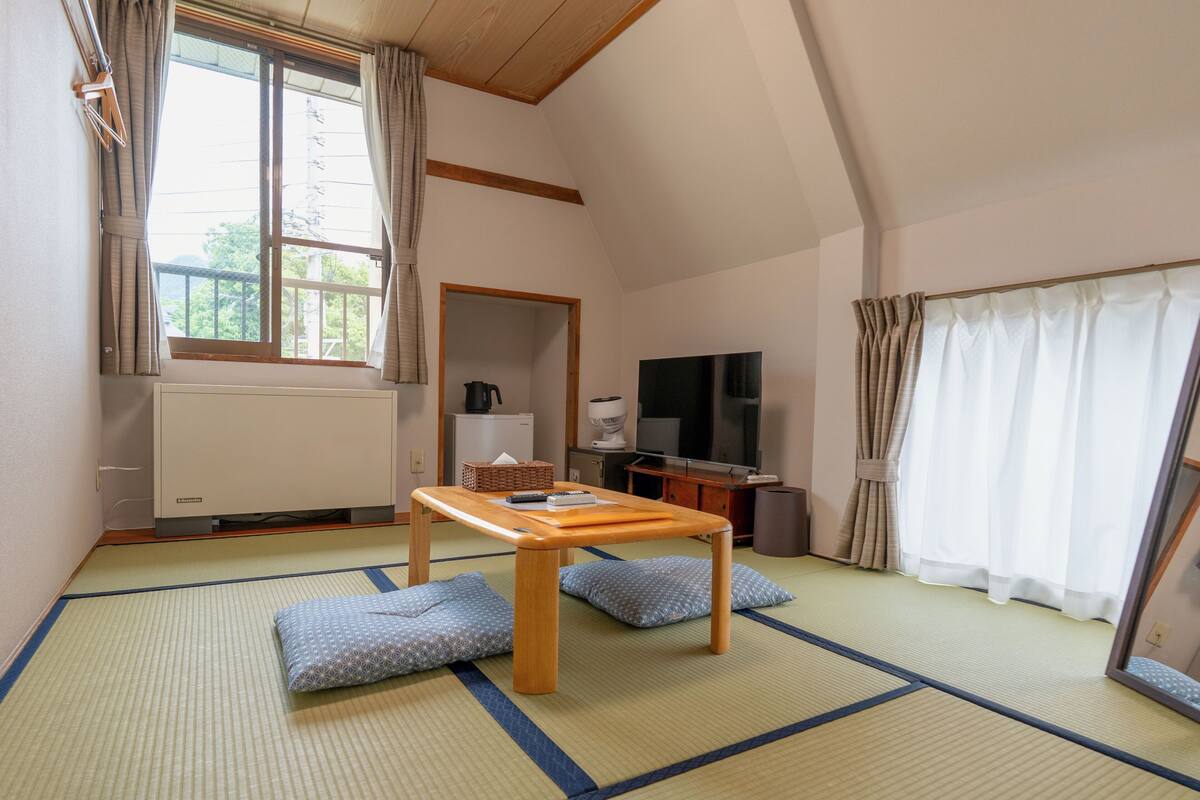 A traditional Japanese room with tatami flooring, a low wooden table, cushions, a television, and a window with curtains.
