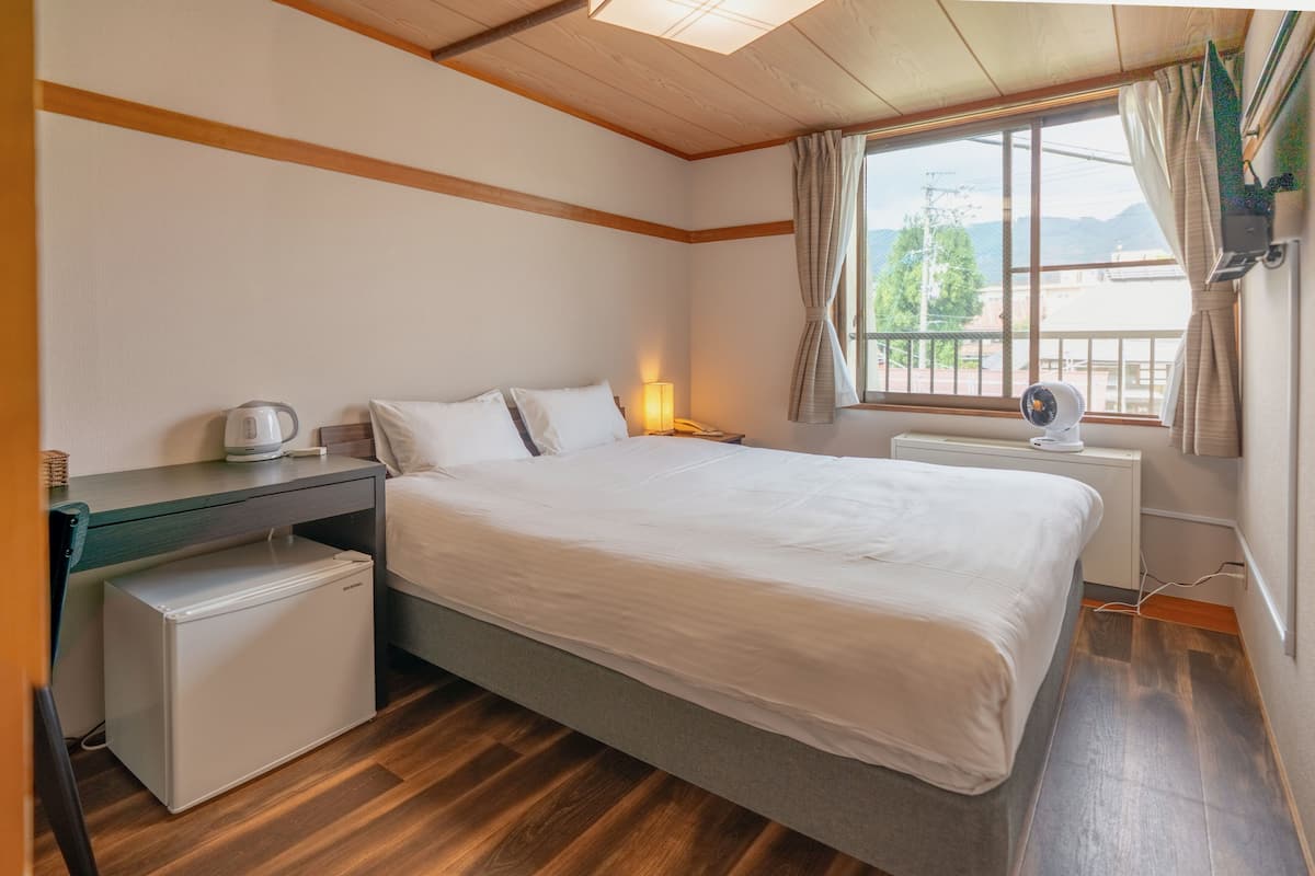 A bedroom with a bed, a desk, a small refrigerator, a lamp, and a window with curtains.