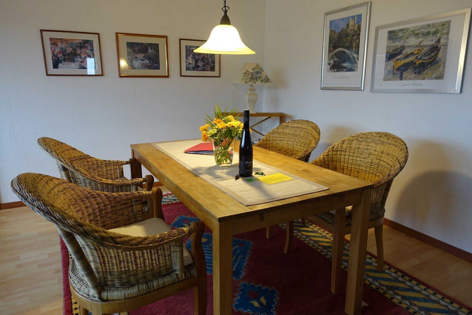 A dining area with a wooden table, wicker chairs, a vase with flowers, and framed pictures on the wall.