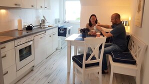 Moderni keittiö, jossa on valkoiset kaapit, sisäänrakennettu uuni sekä ruokailutila, jossa on valkoinen pöytä ja tuolit.