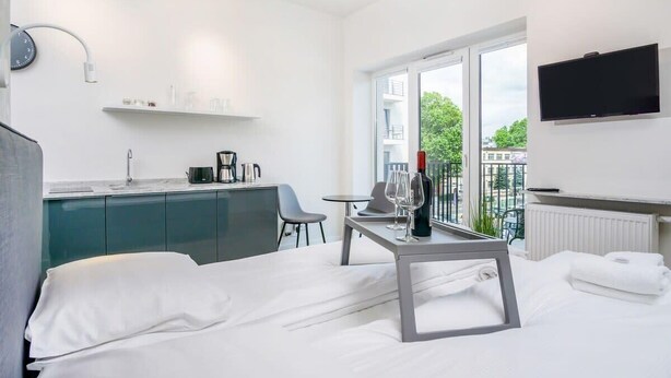 A modern hotel room with a bed, a small table with a wine glass, a TV, and a balcony with a view of the city. - Stylish Studio in the Center of Poznań | Balcony | Close to MTP (Poznan)