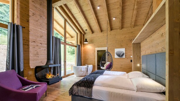 Un dormitorio de cabaña de madera moderno con una cama grande, una chimenea, un sillón colgante y vistas al exterior.