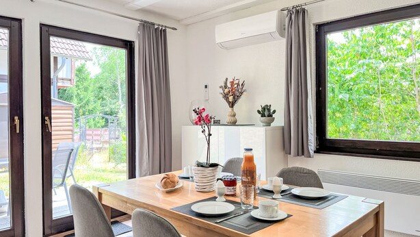 Un coin repas avec une table en bois dressée pour quatre personnes, une plante en pot et une vue sur la verdure à l’extérieur.