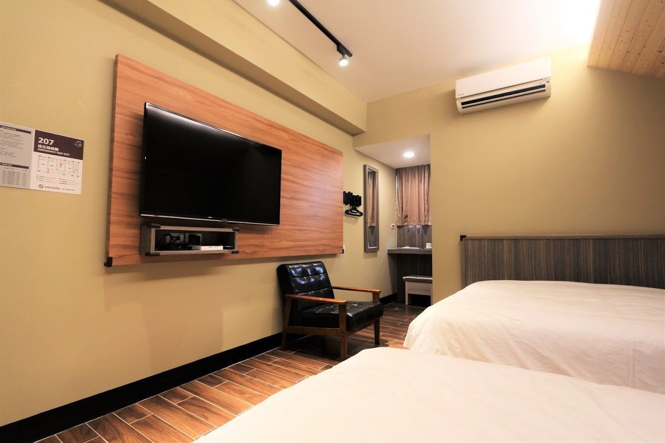 A hotel room with a flat-screen TV mounted on the wall, a wooden chair, a bed, and an air conditioning unit.
