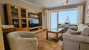 A living room with a television, wooden cabinet, two grey sofas, a glass coffee table, and a view of the sea through large windows.