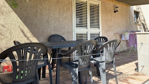 Um pátio externo com mesa e cadeiras, uma janela com persianas e um aplique de luz fixado na parede.