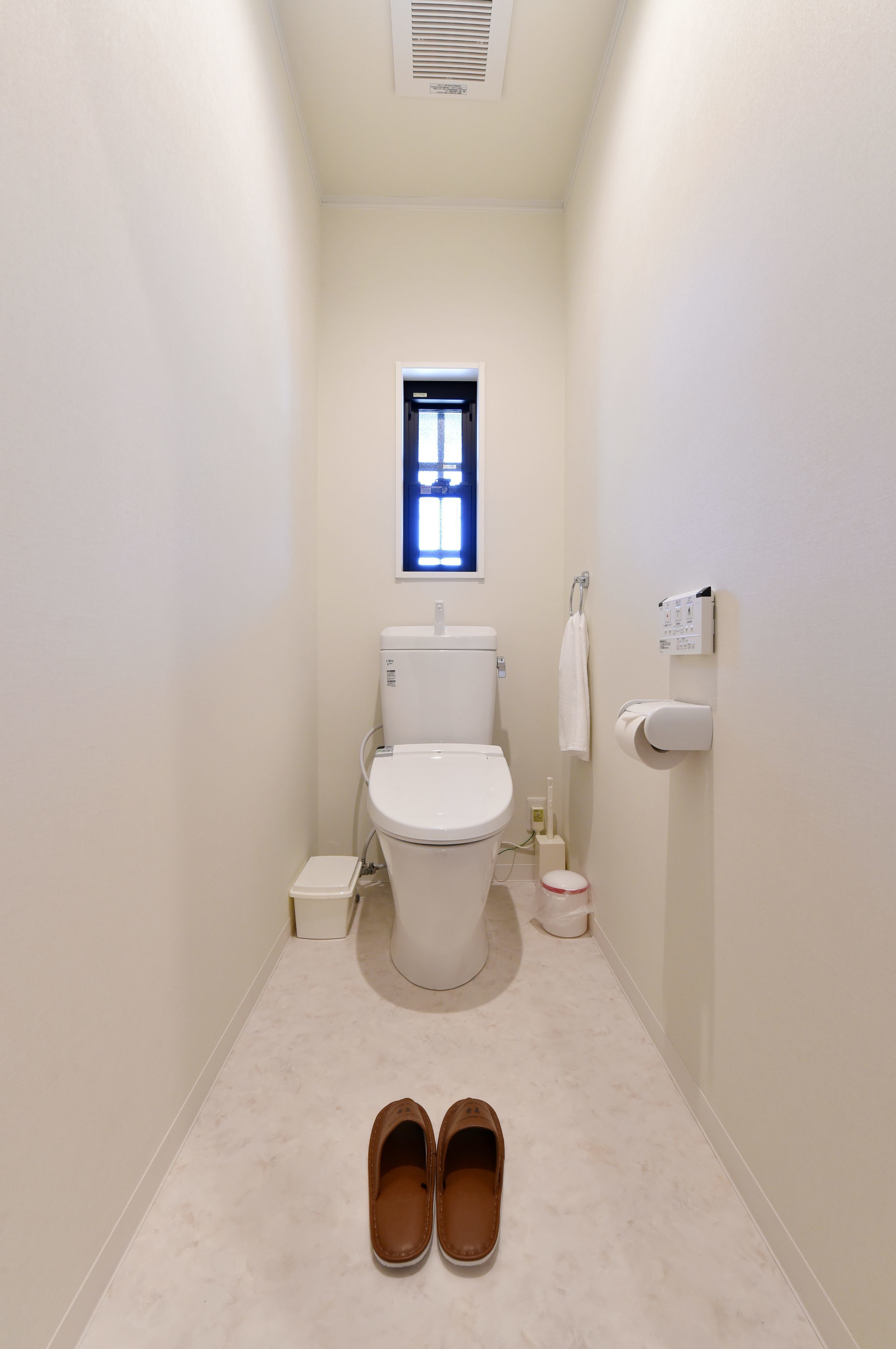 Casa de banho estreita com sanita, uma pequena janela, um porta-toalhas e um par de chinelos no chão.