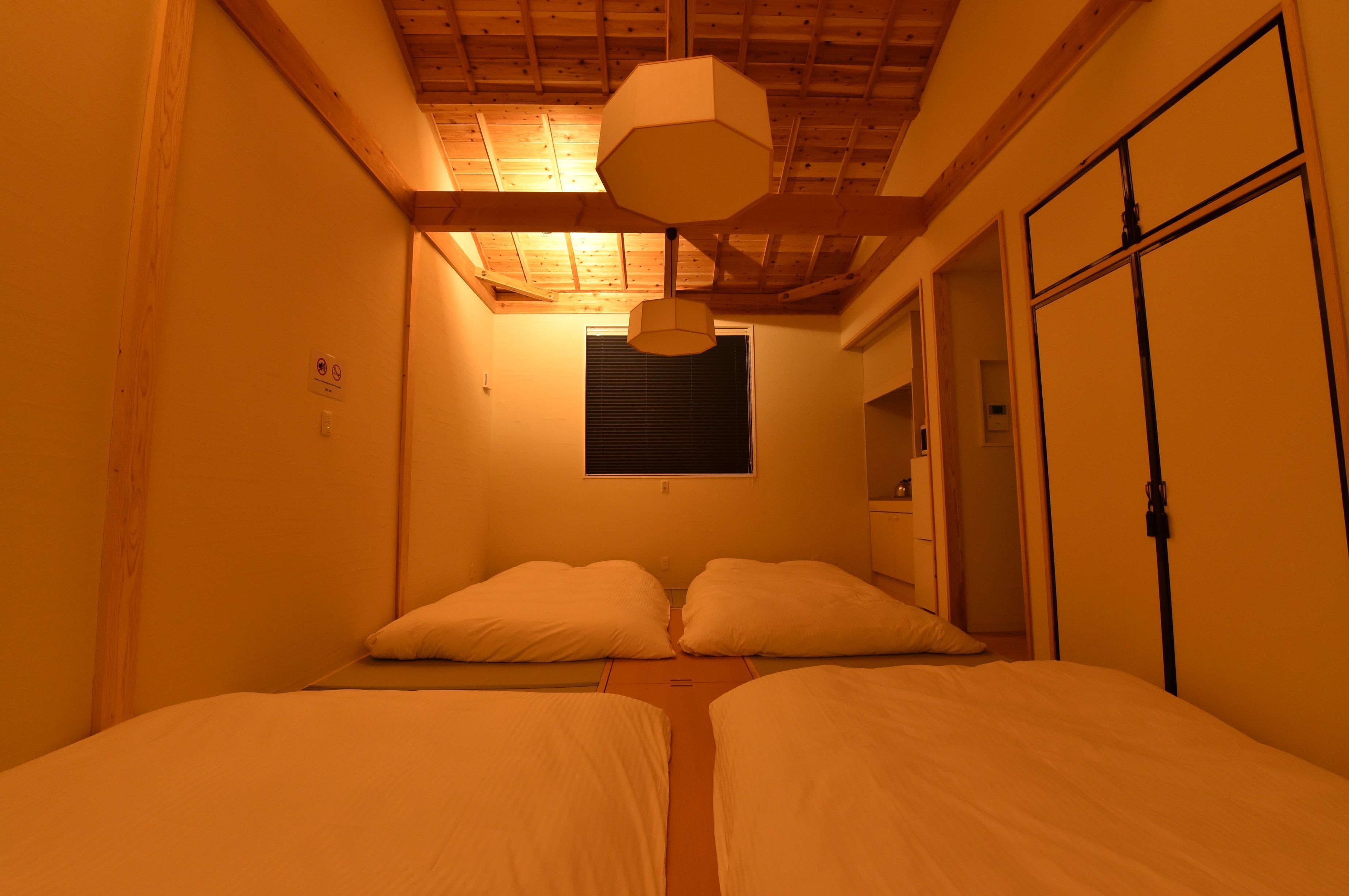 A room with two beds, a wooden ceiling, and a window with blinds.