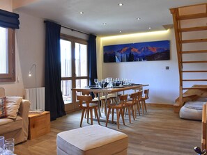 Un comedor con una mesa dispuesta para seis personas, sillas de madera, un sofá, un pequeño reposapiés y un ventanal con cortinas.
