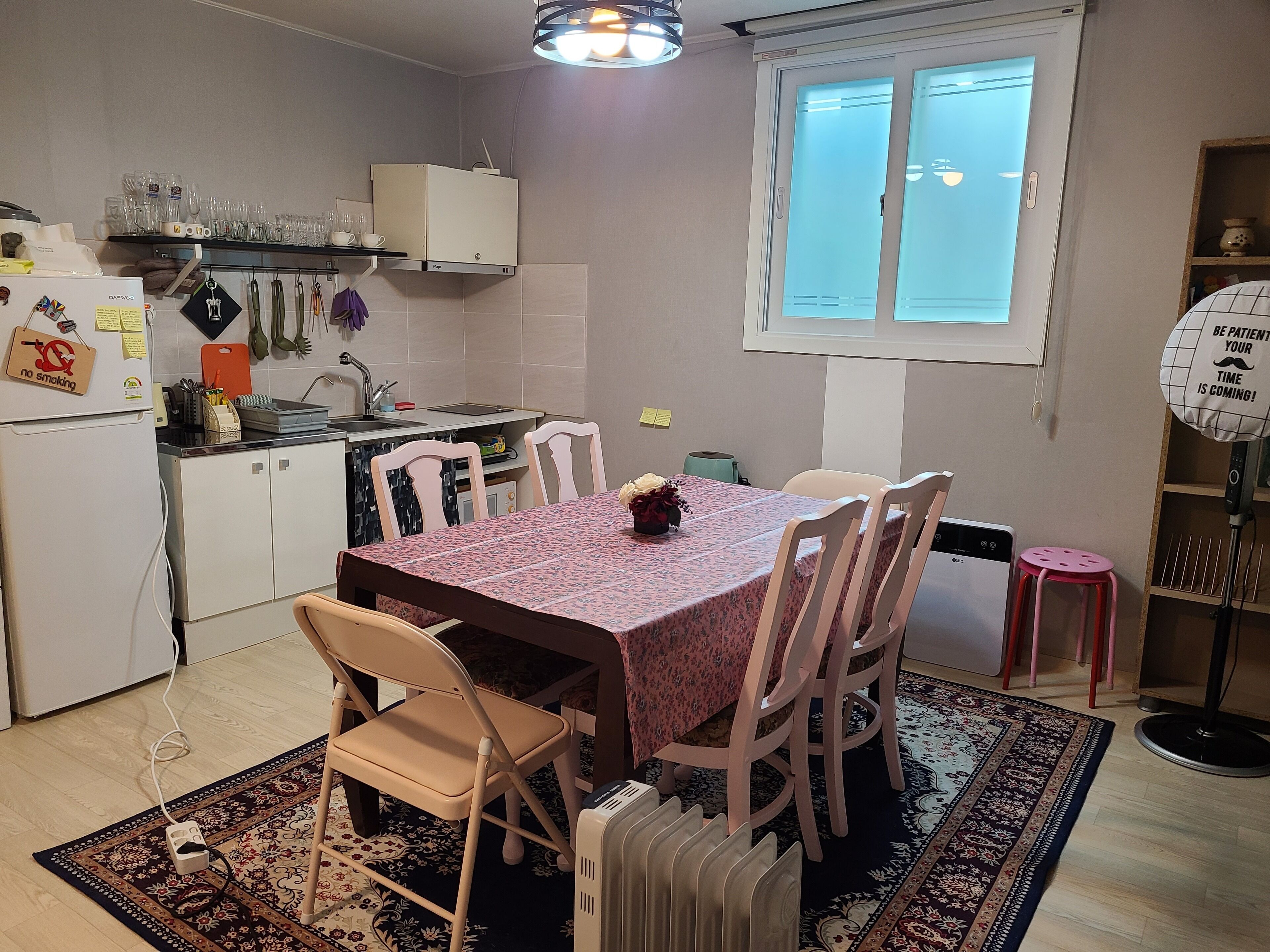 A compact kitchen with a dining table, chairs, a small stool, a radiator, and a window with blinds.