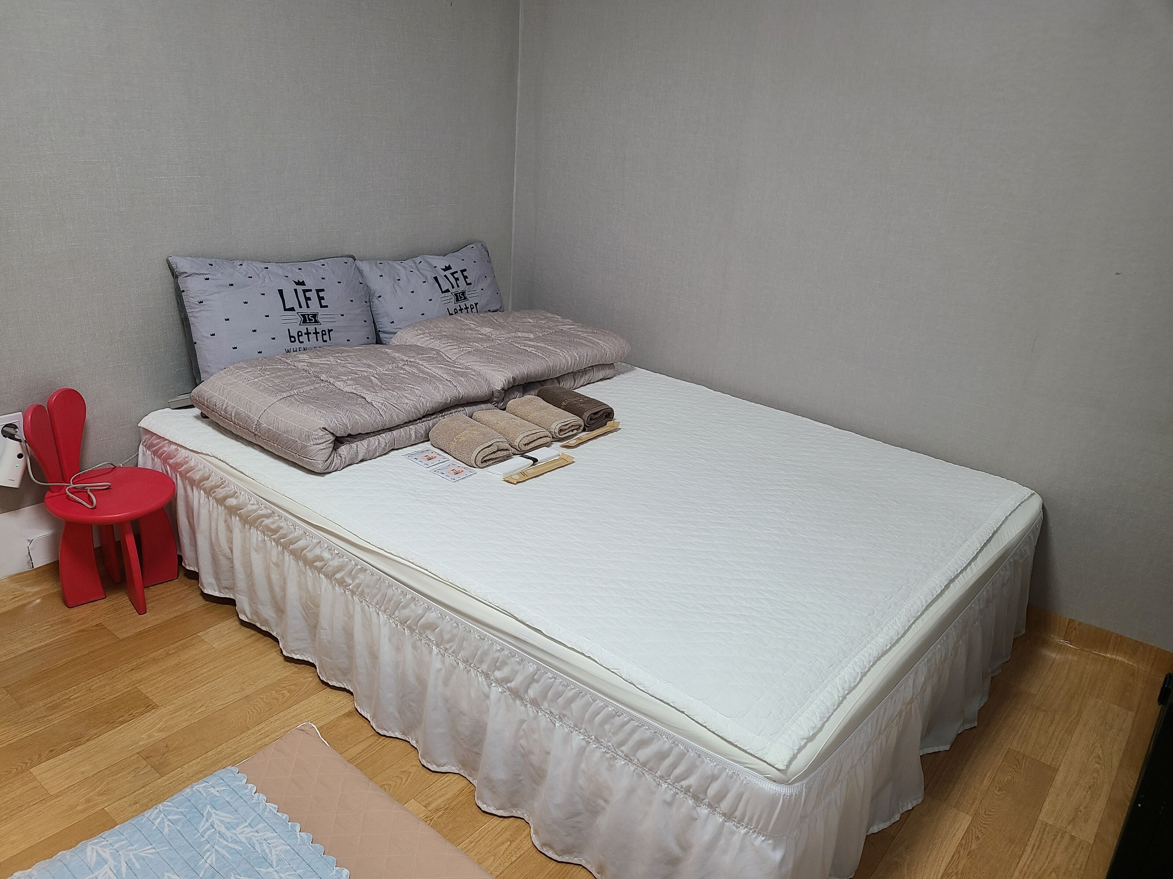 Una cama bien tendida con un edredón blanco y una almohada decorativa, una silla roja y una alfombra pequeña sobre piso de madera.