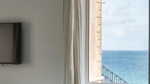 A bedroom with a bed, a TV mounted on the wall, and a window with a view of the sea.