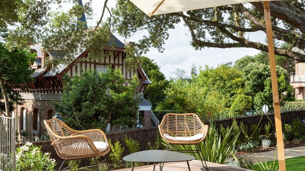 An outdoor patio with wicker chairs and a table, surrounded by lush greenery and a historic building in the background.