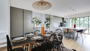 A modern dining area with a black table set for six, surrounded by black chairs. There is a large window providing natural light, and a kitchen area is visible in the background.