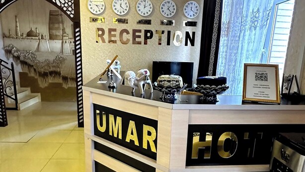 A reception desk at the Umar Hotel with clocks displaying different cities, a framed certificate, and a minibar. - Hotel UMAR (Khiva)