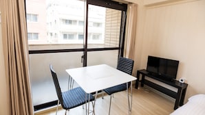 Habitación de hotel con puerta corrediza de vidrio, una mesa blanca con dos sillas, un mueble negro para televisor y una cama.
