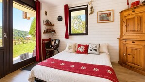Een slaapkamer met een bed, een houten kledingkast, een bureau met stoel, een raam met gordijnen, en uitzicht op groen.