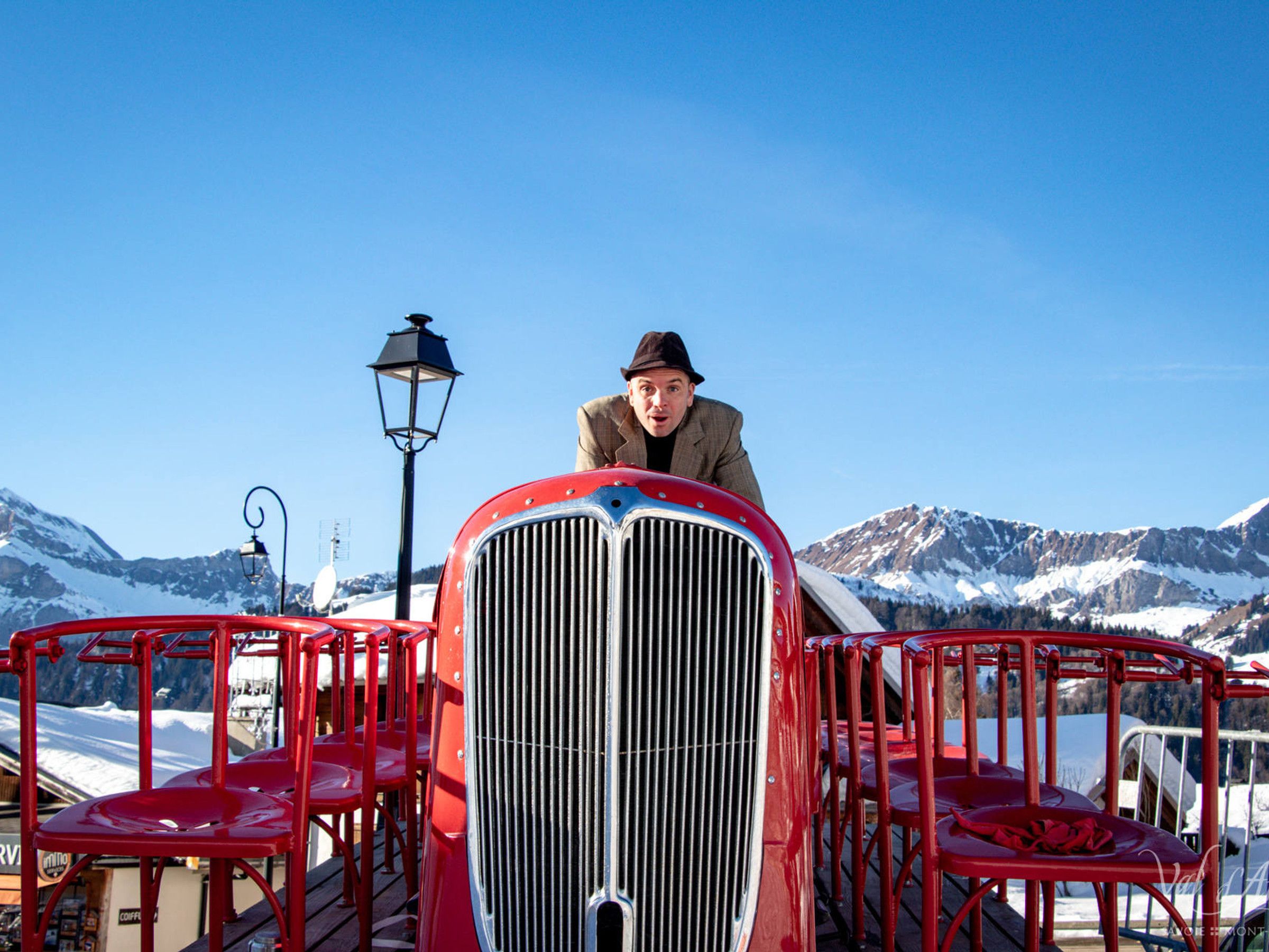 A vintage car with a prominent grille, surrounded by red chairs, set against a mountainous backdrop.