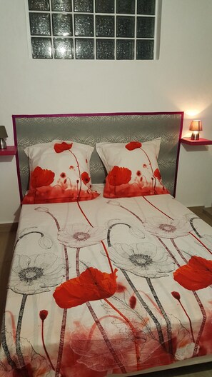Cozy bed with a floral-patterned bedspread, situated against a wall with a window above, and a bedside table with a lamp.
