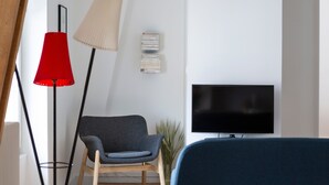 Ein modernes Wohnzimmer mit Fernseher, roter Stehlampe, grauem Sessel und Bücherregal.
