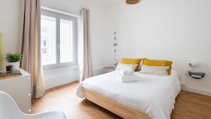 Ein Schlafzimmer mit einem hölzernen Bett, weißer Bettwäsche, einem Nachttisch mit Lampe, einem kleinen Schreibtisch mit Pflanze und einem Fenster mit Vorhängen.
